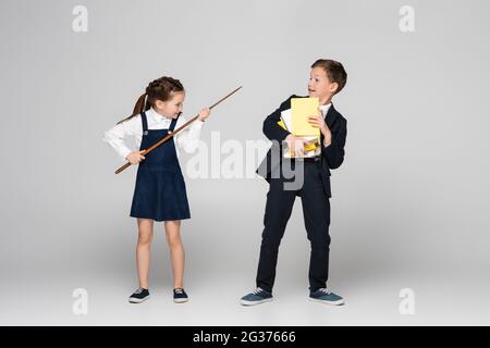 sorridente studentessa che tiene il bastone di puntamento e minaccia scolaro in uniforme con pila di libri su grigio Foto Stock