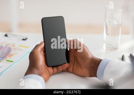 Irriconoscibile medico maschio nero utilizzando smartphone con schermo vuoto, consultando i pazienti, dando consigli a distanza, raccolto Foto Stock