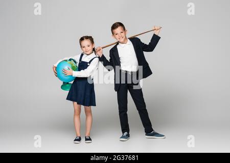 felice scolaro che tiene il bastone di puntamento vicino alla ragazza sorridente in abito con il globo su grigio Foto Stock