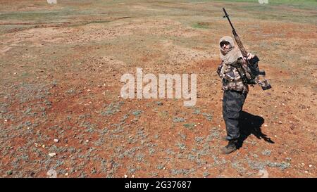 Cecchino soldato nel deserto. Si alza, guarda la macchina fotografica e mostra il suo fucile. Il concetto di conflitti militari. Foto Stock