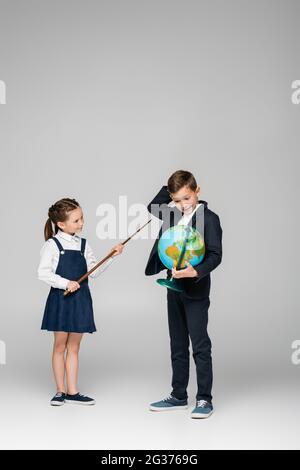 sorridente studentessa tenendo puntato bastone vicino confuso ragazzo con globo su grigio Foto Stock