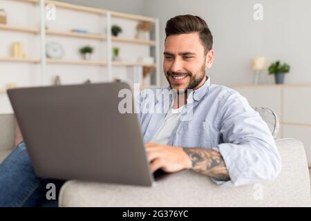 Stile di vita professionale moderno. Felice uomo tatuato seduto sul divano a casa e lavorando su un computer portatile Foto Stock