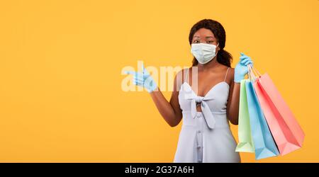 Giovane donna nera che tiene borse regalo, indossando maschera facciale e guanti di gomma, puntando allo spazio vuoto su sfondo arancione Foto Stock