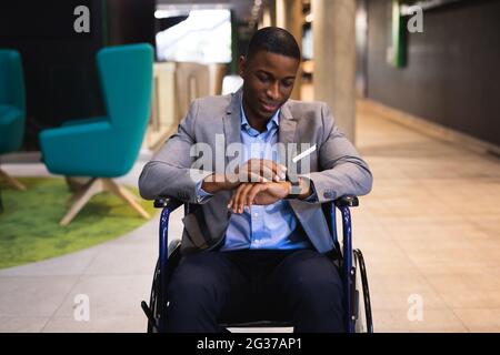 Uomo d'affari afro-americano disabile che usa lo smartwatch mentre si siede su sedia a rotelle in un ufficio moderno Foto Stock