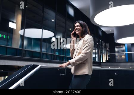 Donna d'affari caucasica sorridente mentre parla su smartphone in piedi vicino alle scale in ufficio Foto Stock