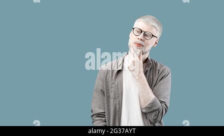 Ragazzo albino pensieroso con aspetto insolito che tocca il mento e pensando alla domanda, sfondo blu, panorama Foto Stock