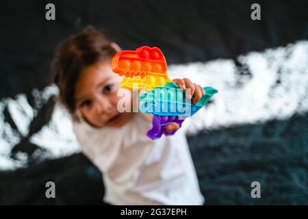 Bambina con giocattolo popit in colori arcobaleno. Giocattolo in silicone a forma di dino per ridurre lo stress. Bolle sensoriali fidget trendy. Bambino che gioca a casa Foto Stock