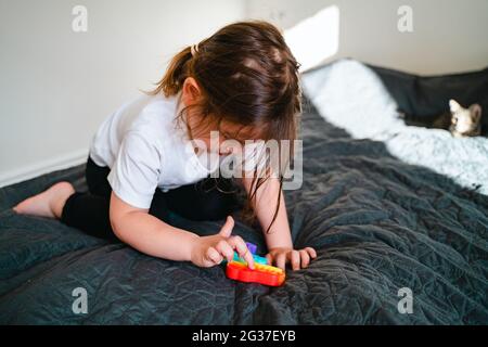 Bambina con giocattolo popit in colori arcobaleno. Giocattolo in silicone a forma di dino per ridurre lo stress. Bolle sensoriali fidget trendy. Bambino che gioca a casa Foto Stock