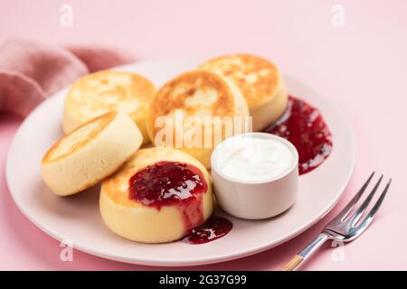 Syrniki, frittelle di formaggio cottage con marmellata di lamponi e panna acida su un piatto rosa, vista closeup Foto Stock