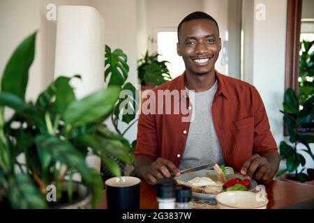 Sorridente giovane africano che mangia la colazione al suo tavolo da pranzo Foto Stock