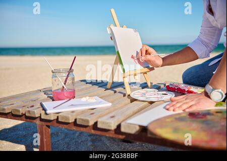 Immagine ritagliata di un artista che tiene il pennello e la barca di pittura con acquerelli, seduto su una chaise lounge in legno con sfondo mare Foto Stock
