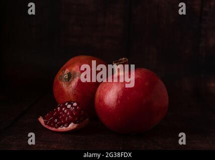 Melograni maturi succosi di colore rosso scuro su sfondo scuro con un piccolo pezzo di melograno spezzato con grani di frutta. Foto Stock