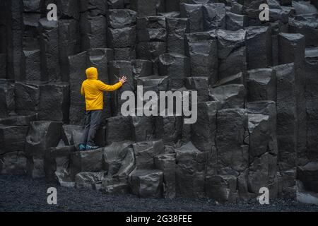 Uomo adulto in giacca gialla sulle colonne di basalto sulla spiaggia di sabbia nera di Reynisfjara in Islanda Foto Stock