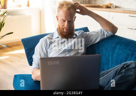 Sorridente giovane uomo rosso trascorre il tempo libero in rete sdraiato sul divano di casa, utilizzando un computer portatile per guardare film, chattare e inviare messaggi online Foto Stock