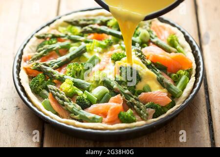 Preparazione di quiche con salmone, broccoli e asparagi verdi, ricoperti di latte e uova. Crostata grezza - forno pronto. Foto Stock
