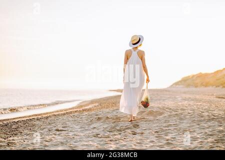 Concetto di stile di vita riciclabile riutilizzabile. Donna in abiti naturali camminare lungo una spiaggia e tenere riutilizzabile mesh borsa della spesa con verdure Foto Stock