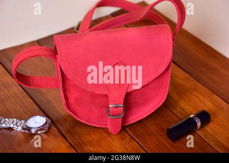 Borsa rossa in pelle da donna alla moda su sfondo di legno, accanto a lei un orologio e rossetto Foto Stock