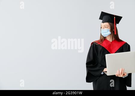 Studentessa laureata in maschera protettiva e con laptop su sfondo chiaro Foto Stock