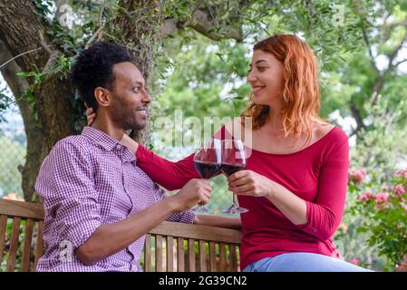 dolce coppia multietnica gustando un bicchiere di vino insieme all'aperto in un giardino seduto su una panchina. amici felici e diversi che si divertono a bere alcolici Foto Stock