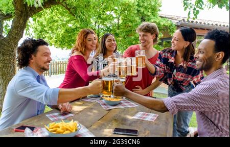 gruppo multietnico di persone che si gustano la birra insieme all'aperto seduto su un tavolo da giardino. amici felici e diversi divertirsi a fare un brindisi bere alcol Foto Stock