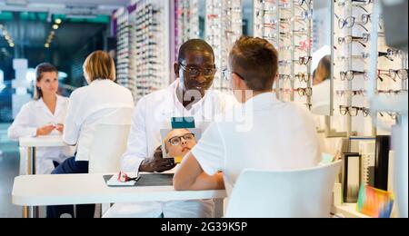 Sorridente afro-americano oftalmologo aiutare ragazzo teen a scegliere gli occhiali in negozio ottico, mostrando specchio Foto Stock