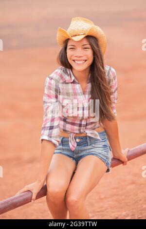 Cowgirl Texas donna sorridente felice sul ranch fattoria di campagna che indossa cappello cowboy, camicia occidentale e jeans shorts. Giovane ranch asiatico-americano multirazziale Foto Stock