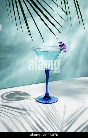 Cocktail di colore blu sul tavolo con sole ombre e riflesso d'acqua. Su sfondo blu con foglie di palma, vista frontale. Foto Stock