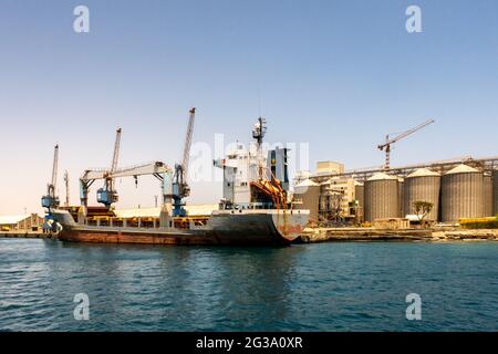 Nave di grandi dimensioni che viene caricata con carico nel porto di Port Sudan. Enormi gru portuali e silos di grano a file sullo sfondo. Foto Stock