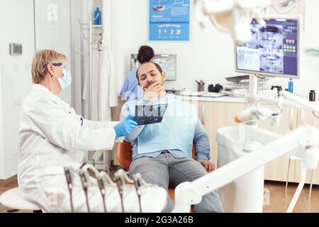 Dentista donna anziana che esamina la radiografia del dente con l'uomo malato che discute il mal di denti durante l'appuntamento stomatologico. Paziente seduto su sedia dentale in ospedale odontoiatria ufficio Foto Stock