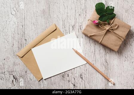 Biglietto d'auguri bianco bianco per mockup o invito, confezione regalo avvolta con carta kraft e germoglio rosa sul tavolo grigio. Foglio di carta vuoto per il testo Foto Stock