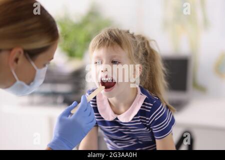 Otorhinolaringologo medico in maschera medica protettiva che esamina la gola della bambina con spatola in legno Foto Stock