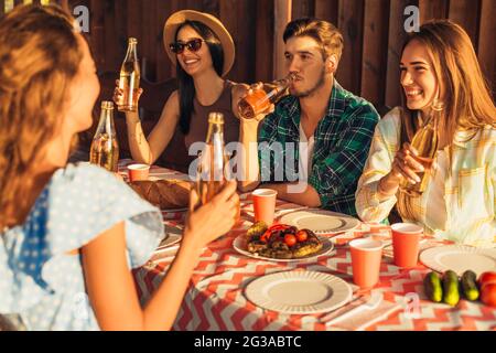 I giovani amici si divertono, bevendo bevande a casa cena all'aperto, gente felice mangiare barbecue insieme in natura, al tramonto Foto Stock