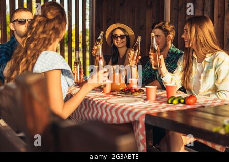 I giovani amici si divertono, bevendo bevande a casa cena all'aperto, gente felice mangiare barbecue insieme in natura, al tramonto Foto Stock