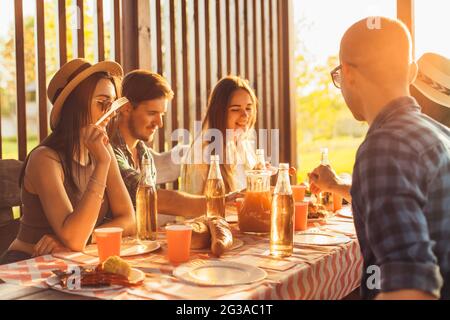 I giovani si divertono sulla terrazza, bevono bevande e chiacchierano mentre i loro amici servono carne alla griglia all'aperto al tramonto Foto Stock