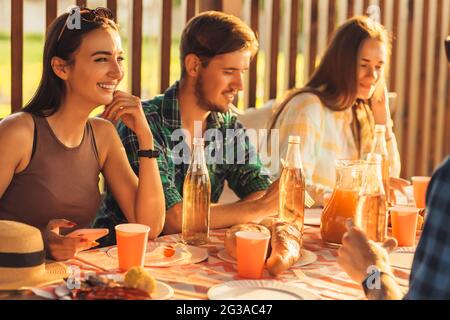 I giovani amici si divertono, bevendo bevande a casa cena all'aperto, gente felice mangiare barbecue insieme in natura, al tramonto Foto Stock