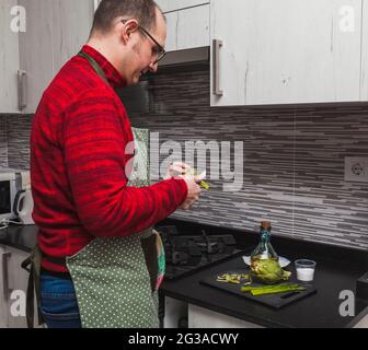 Un uomo in un vello rosso e un grembiule verde che cucinano carciofi e asparagi alla griglia in cucina. Sta tagliando i carciofi. Foto Stock