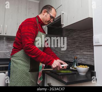 Un uomo in un vello rosso e un grembiule verde che cucinano carciofi e asparagi alla griglia in cucina. Sta tagliando asparagi. Foto Stock