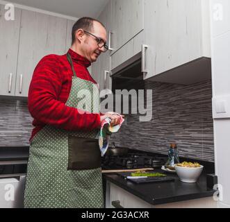 Un uomo in un vello rosso e un grembiule verde che cucinano carciofi e asparagi alla griglia in cucina. Sta asciugando le mani. Foto Stock