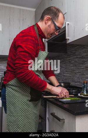 Un uomo in un vello rosso e un grembiule verde che cucinano carciofi e asparagi alla griglia in cucina. Sta tagliando asparagi. Foto Stock