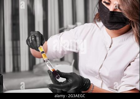 Primo piano di una giovane donna scienziata in maschera medica che tiene la provetta con il campione di sangue. Tecnico femminile in guanti sterili con siringa e preparazione del materiale per il processo di plasmolifting. Foto Stock