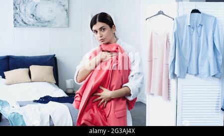 giovane donna in accappatoio che tiene il blazer rosa vicino al letto con i vestiti in camera da letto Foto Stock