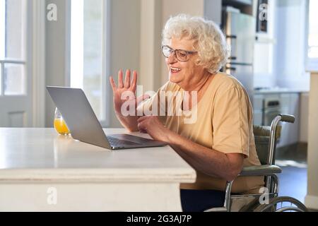 Donna anziana felice in sedia a rotelle che fa la videochiamata con la famiglia a casa e che agita Foto Stock