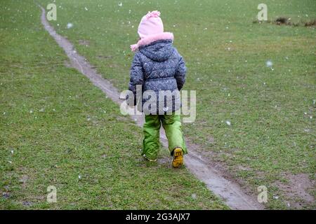 Vista posteriore della bambina di 5 anni in abiti caldi che cammina lungo uno stretto sentiero fangoso su un prato in inverno durante la nevicata. Visto in Germania a Janua Foto Stock