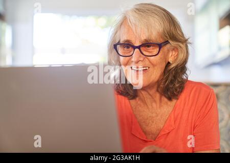 Sorridente donna anziana che parla di video o scrive e-mail sul computer portatile a casa Foto Stock