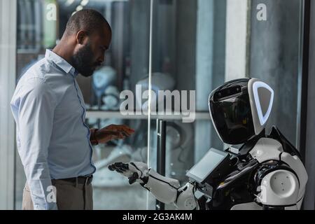 uomo d'affari afroamericano in piedi vicino robot umanoide in ufficio Foto Stock