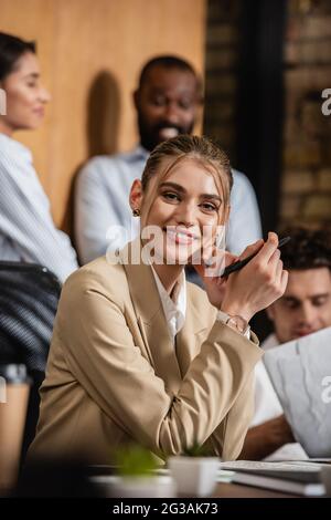 donna d'affari gioiosa che guarda la macchina fotografica vicino i soci interracial offuscati di affari Foto Stock