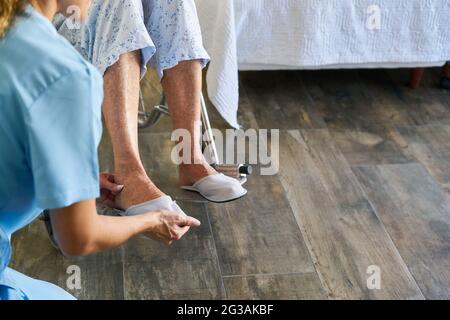 L'infermiere geriatrico aiuta il cittadino anziano fragile in una sedia a rotelle a mettere sulle scarpe nella casa di cura Foto Stock