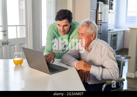 Giovane uomo che aiuta il padre anziano a chattare online sul computer portatile nel suo appartamento Foto Stock