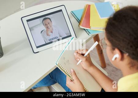 Vista a spalla di una ragazza mista in auricolari che prende appunti nel libro degli esercizi mentre si guarda il webinar sul tablet Foto Stock