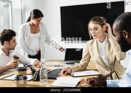 donna d'affari afro-americana incinta che punta al laptop durante la riunione con i colleghi Foto Stock